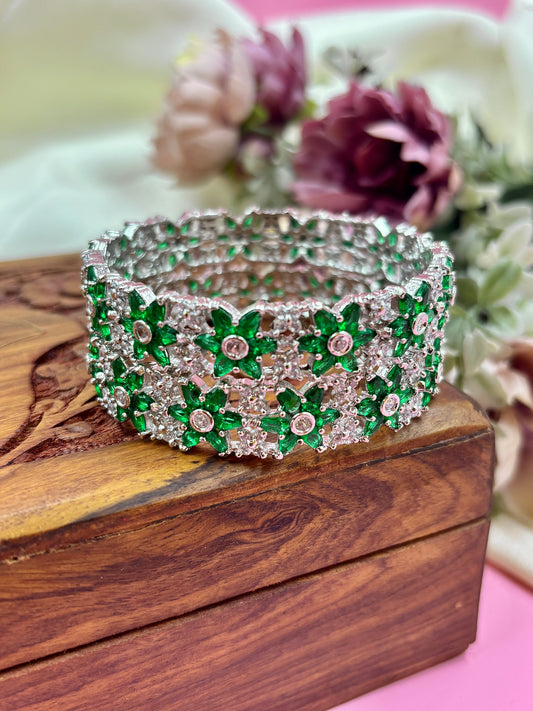 Silver bracelet with green gemstones on a wooden box with flowers in the background by hitesh bangles