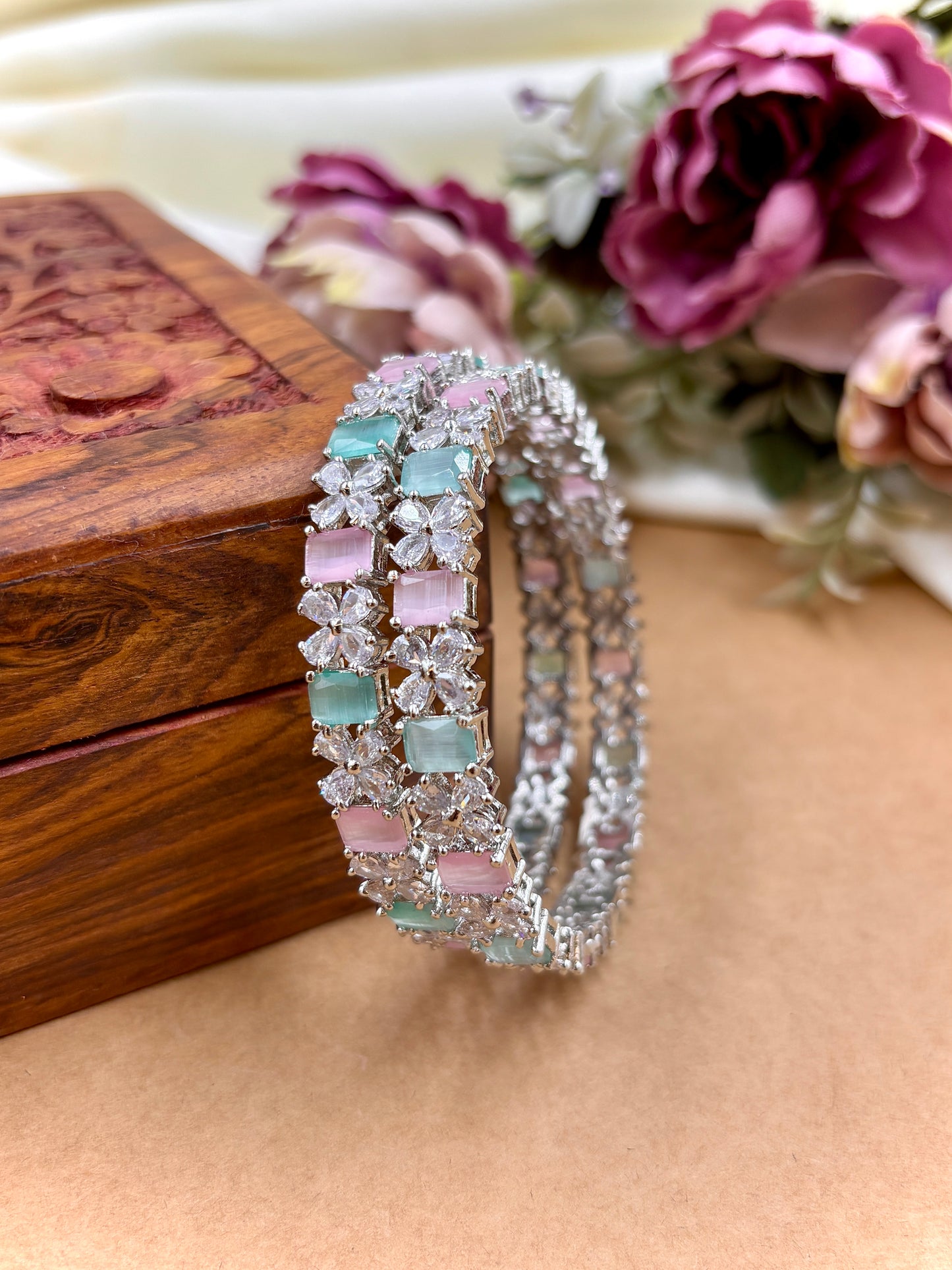 Two decorative bracelets with gemstones on a wooden surface with flowers in the background by hitesh bangles