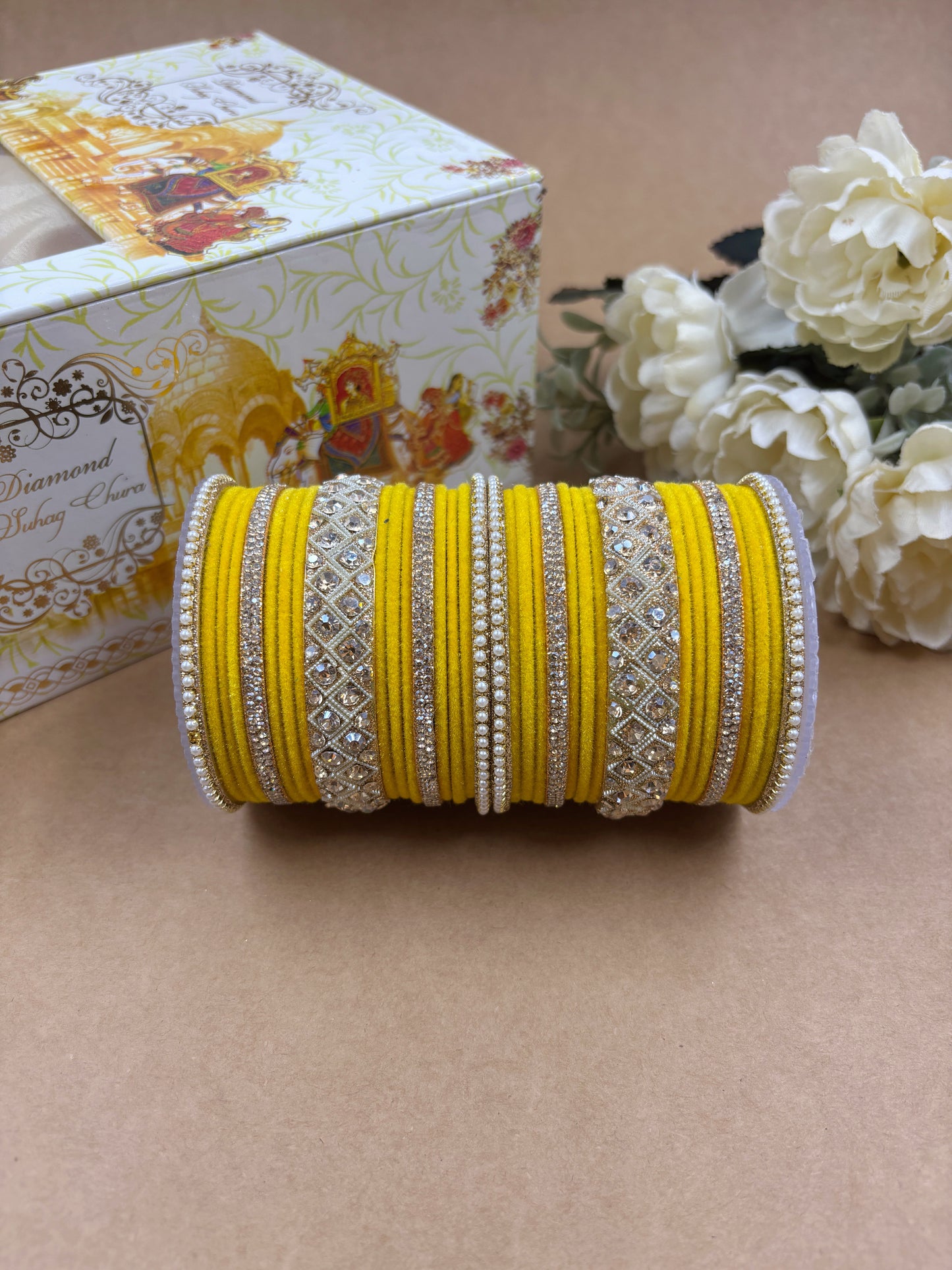 Yellow velvet bridal bangle set featuring intricate gold-toned stonework and pearl detailing, displayed beside a decorative wedding-themed box and white artificial flowers on a brown background by hitesh bangles
