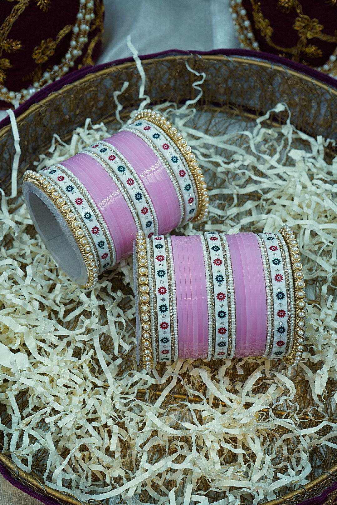 Two pink bangles with gold and multicolor detailing on a white shredded paper background by hitesh bangles