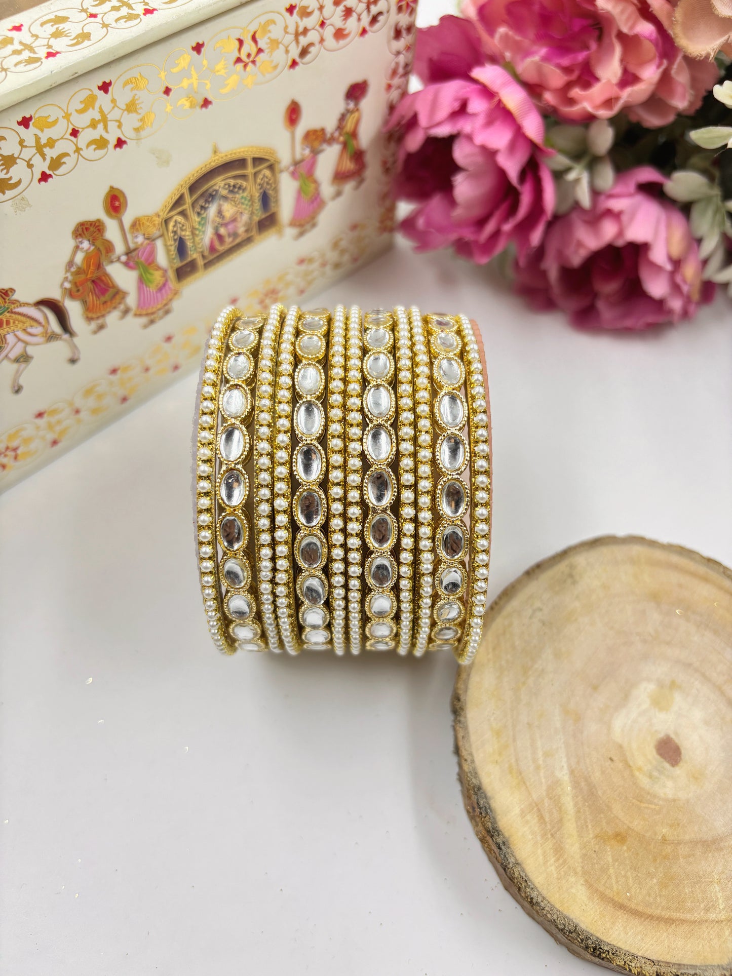 Set of gold and crystal bangles on a white surface with flowers and a decorative box in the background by hitesh bangles