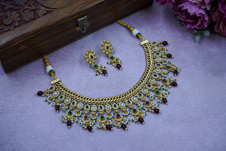 Gold necklace with green and red stones on a purple surface with a wooden box and flowers in the background by hitesh bangles