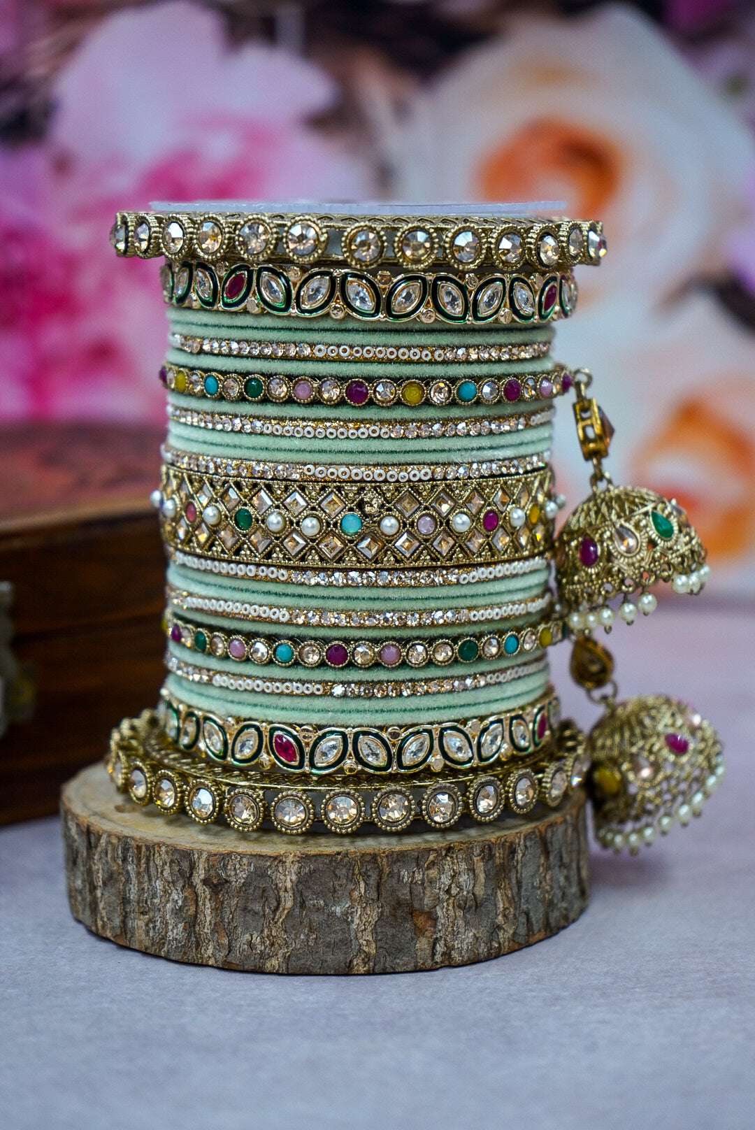 Stack of colorful bangles with a pair of earrings on a wooden block against a blurred floral background by hitesh bangles