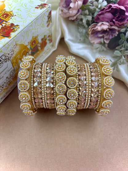 Decorative gold bangles with intricate designs on a beige surface, with a floral arrangement and decorative box in the background by hitesh bangles