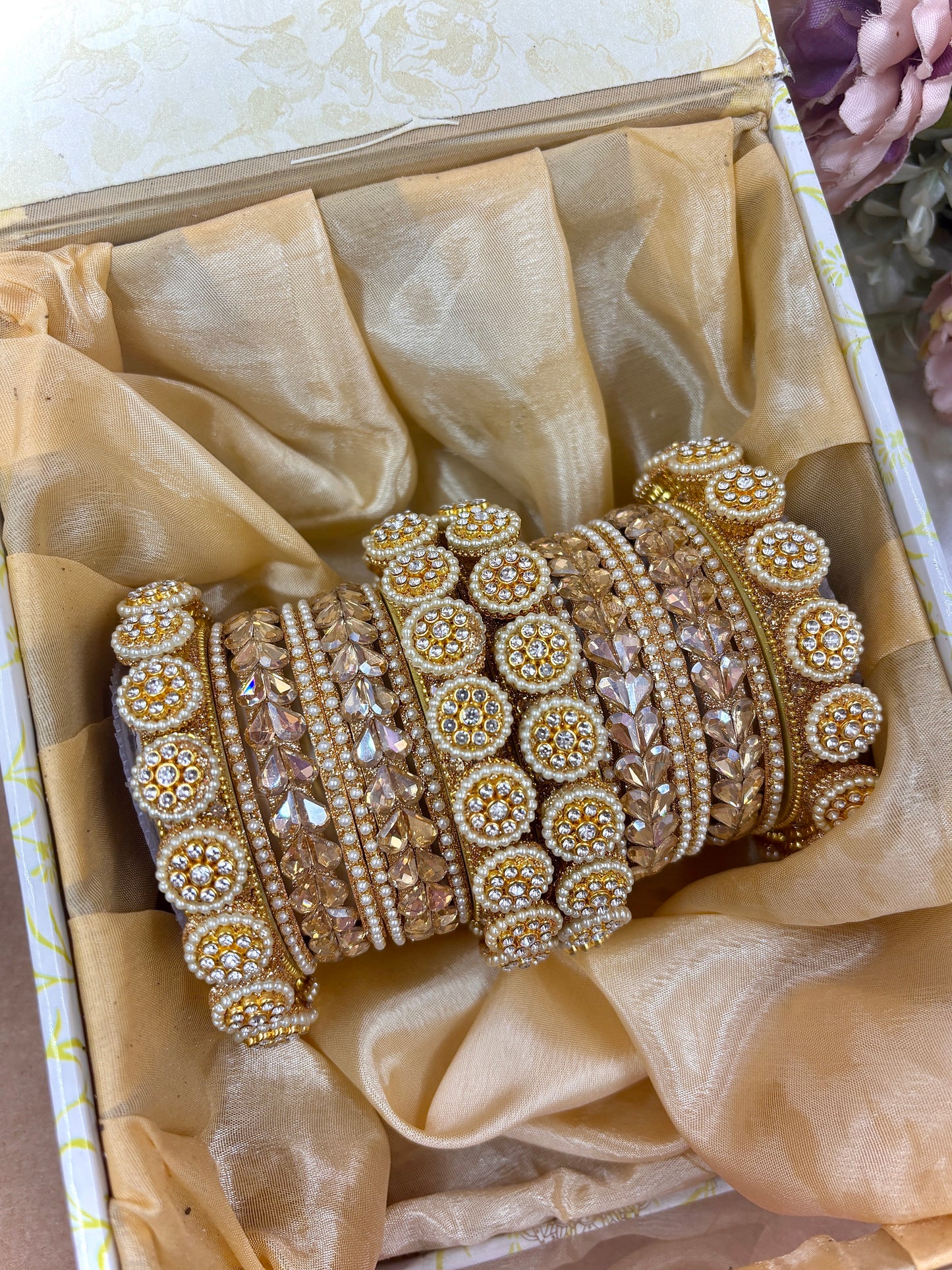 Golden bridal chooda set featuring intricate stone-studded circular kadas and shimmering crystal bead bangles, displayed with floral décor and a decorative box by hitesh bangles
