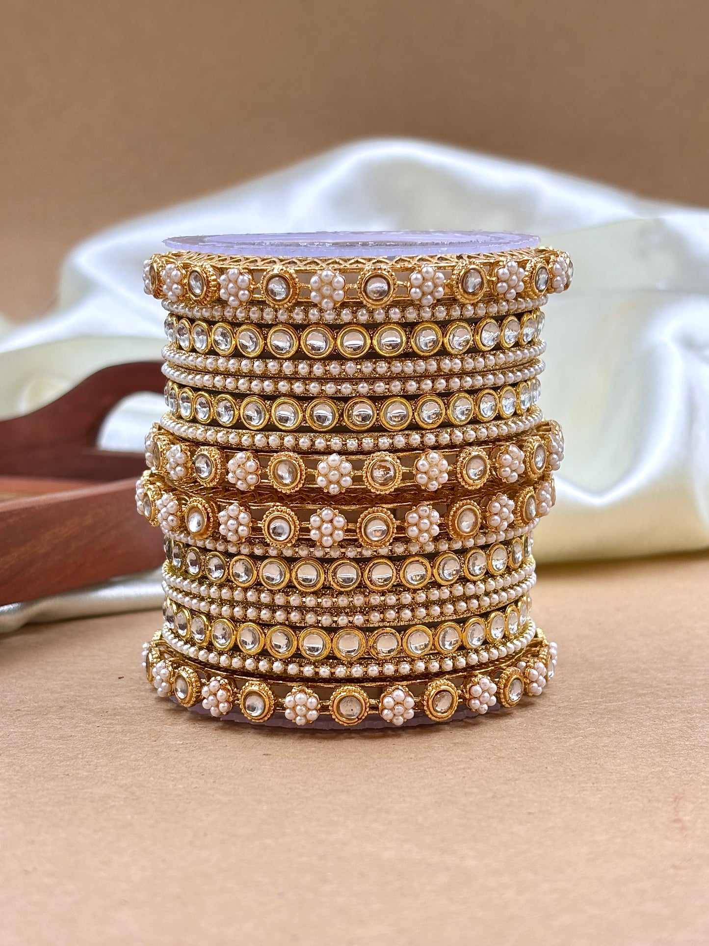 Stack of gold and pearl bangles on a beige surface with a white container in the background by hitesh bangles