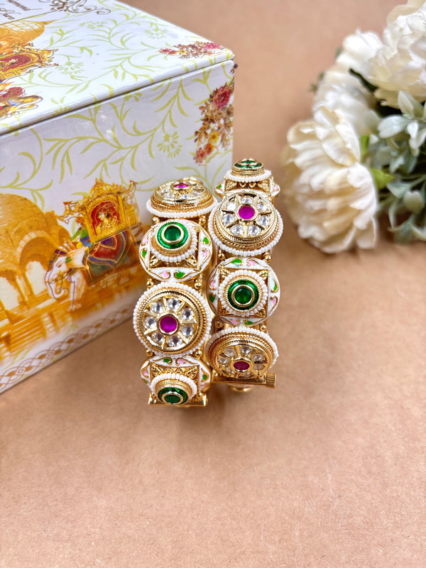 Traditional Kundan Kada with antique gold finish, adorned with intricate circular motifs, ruby red and emerald green stones, placed upright on a beige surface with floral decor and an ornate bridal box in the background.