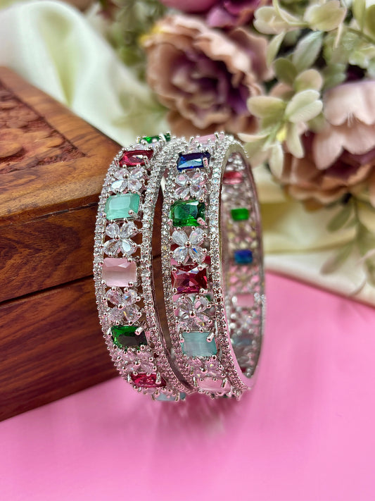 Silver bangles with colorful gemstones on a pink surface with floral background by hitesh bangles