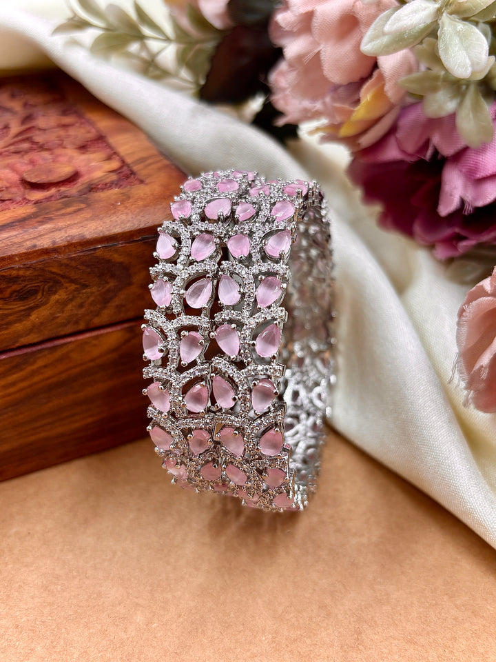 Premium silver-plated kada bangle for women, featuring teardrop-shaped pink American diamonds intricately set in a floral design with sparkling CZ stones, displayed against a soft satin and wooden background with decorative flowers.