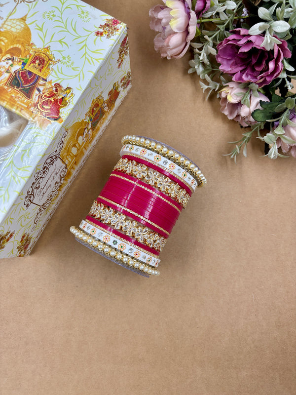 Traditional Rani pink Punjabi bridal chooda set with floral kundan designs, white enamel bangles, and pearl detailing, displayed with floral decor and gift box.