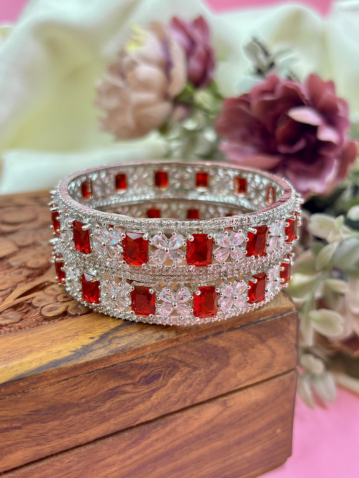 Red Luxe American Diamond Kada for Women – Silver-plated bangle adorned with square-cut red stones and sparkling American diamonds, placed on a carved wooden box with floral background.