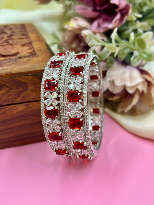 Silver bangles with red gemstones on a pink surface with flowers in the background by hitesh bangles