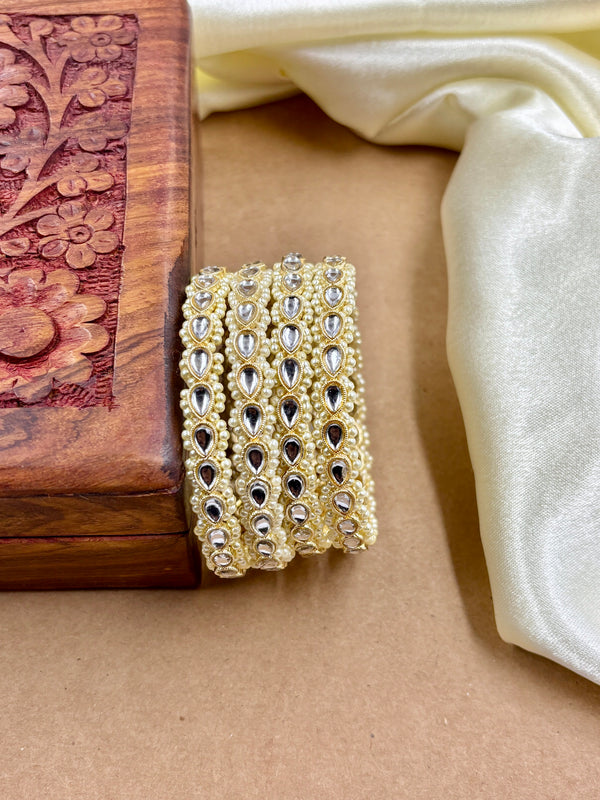 Set of traditional gold bangles adorned with teardrop-shaped kundan stones and intricate pearl detailing, placed beside a carved wooden box and cream satin fabric on a brown background.
