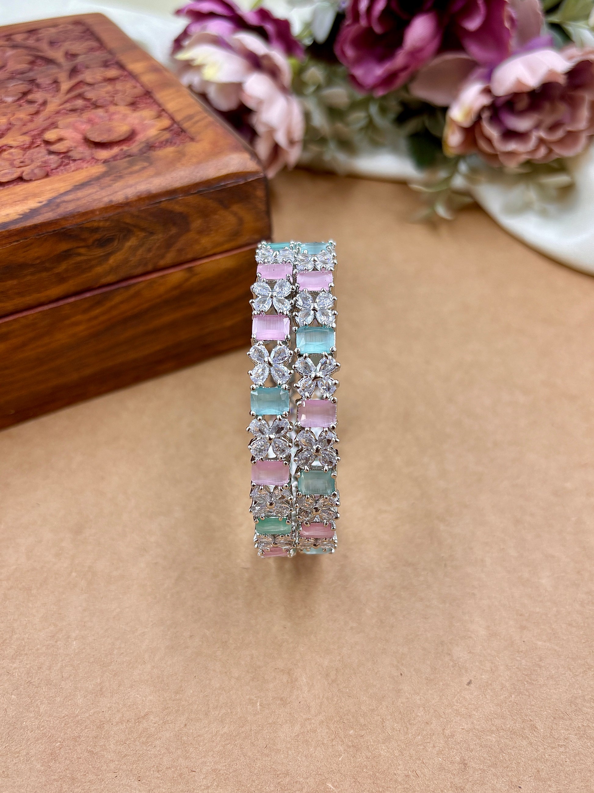 Multicolored gemstone bracelet on a beige surface with a wooden box and flowers in the background by hitesh bangles