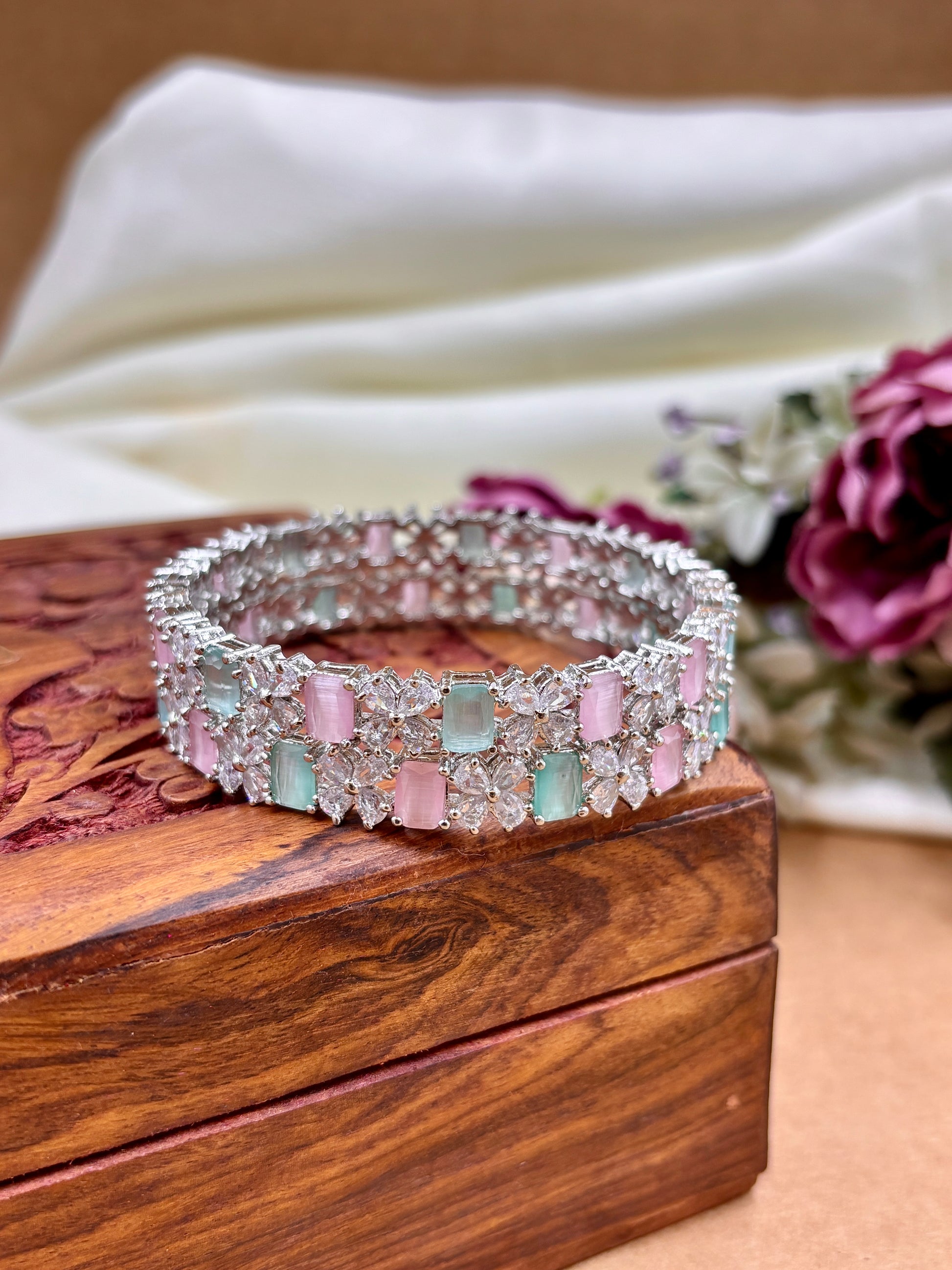 Silver bracelet with pink and green gemstones on a wooden box with flowers in the background by hitesh bangles