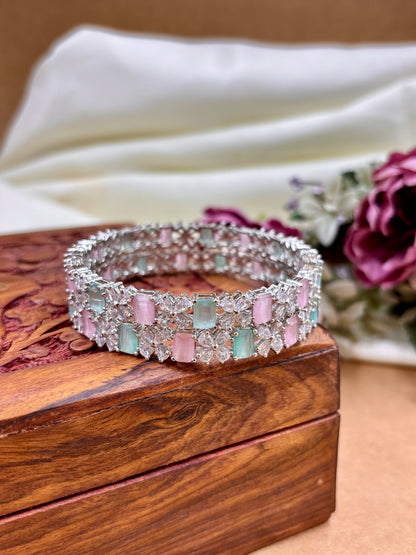 Silver bracelet with pink and green gemstones on a wooden box with flowers in the background by hitesh bangles