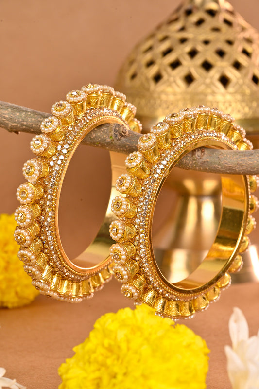 Gold earrings with intricate designs on a wooden branch with yellow flowers and a beige background by hitesh bangles