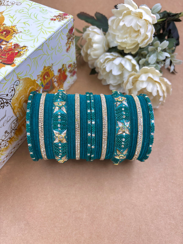 Teal green velvet metal bridal bangle set featuring golden floral kundan designs and shimmering stone detailing, displayed beside white artificial flowers and a decorative wedding-themed box.