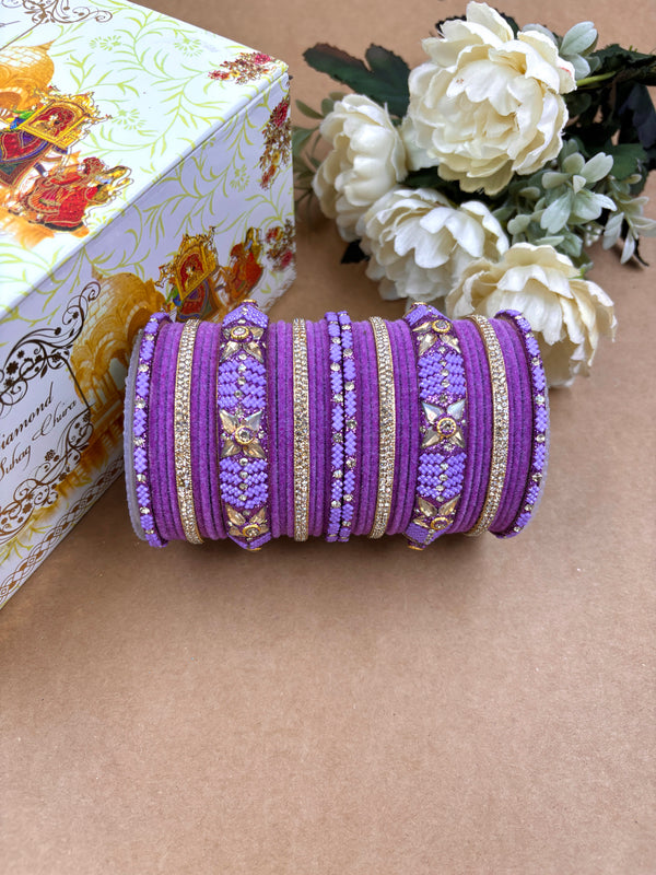 Purple velvet metal bridal bangle set adorned with floral kundan motifs, glittering gold accents, and intricate beadwork, displayed beside white artificial flowers and a decorative wedding-themed box.