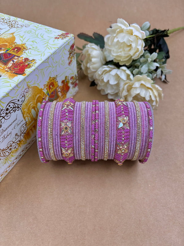 Pink velvet metal bridal bangle set adorned with floral kundan motifs, sparkling stones, and golden accents, styled with white artificial flowers and a decorative wedding box.