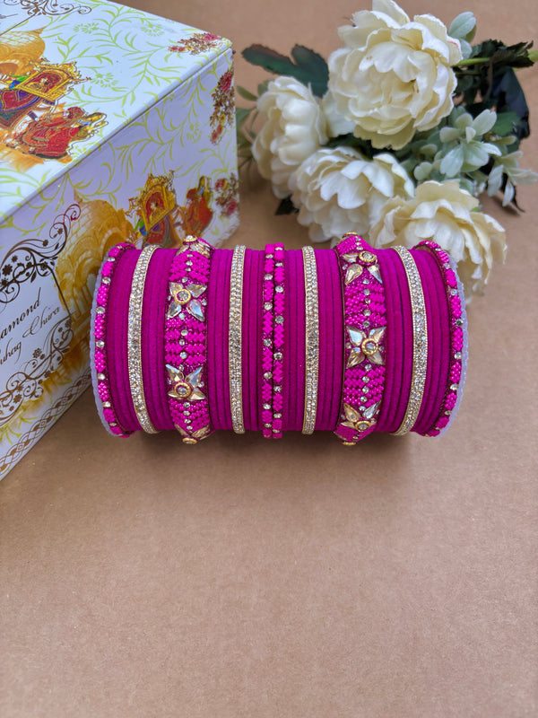 Deep pink metal velvet bangle set for women featuring intricate gold detailing, embedded stones, and floral design motifs Wine Metal Velvet Bangle Set for Women | Elegant and Stylish Wedding Chuda By Hitesh Bangles ideal for bridal wear or festive occasions, displayed with floral décor and designer box in the background.