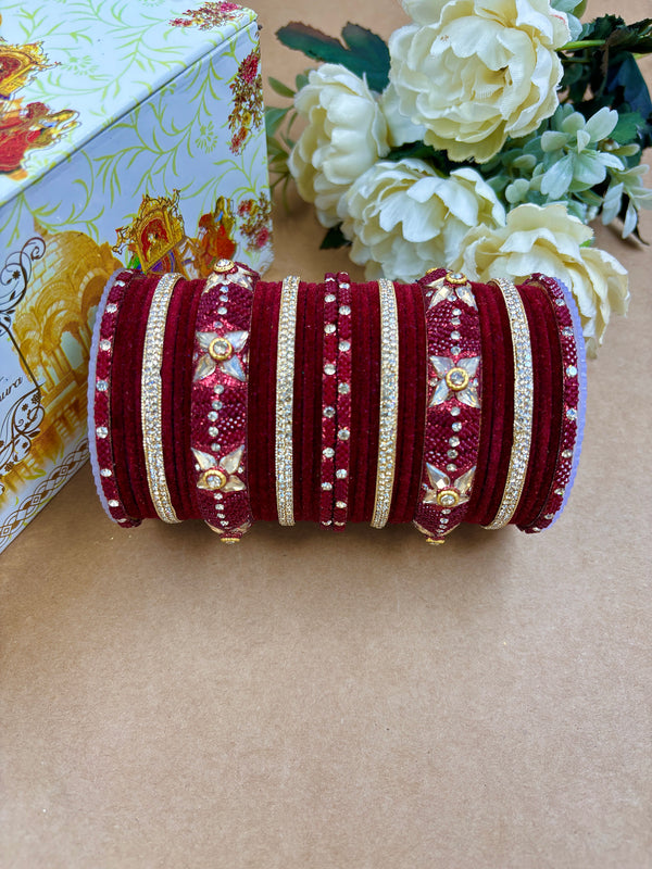 Maroon velvet metal bridal bangle set featuring gold stone detailing, kundan floral motifs, and shimmering accents, styled with white flowers and a designer wedding box.