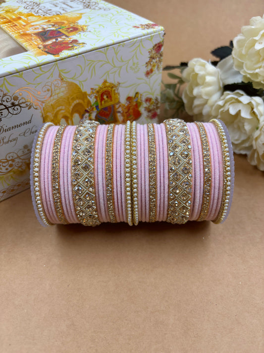 Elegant pink velvet bridal bangle set adorned with intricate gold-toned stone and pearl embellishments, displayed beside a decorative wedding-themed box and white flowers on a brown background by hitesh bangles