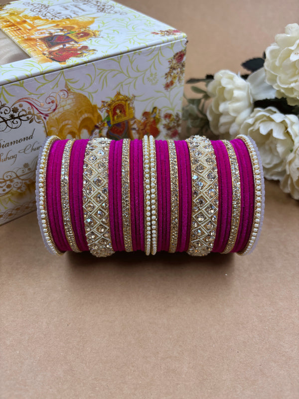Magenta velvet bridal bangle set featuring intricate gold-toned stonework and pearl detailing, displayed beside a decorative wedding-themed box and white artificial flowers on a brown background.