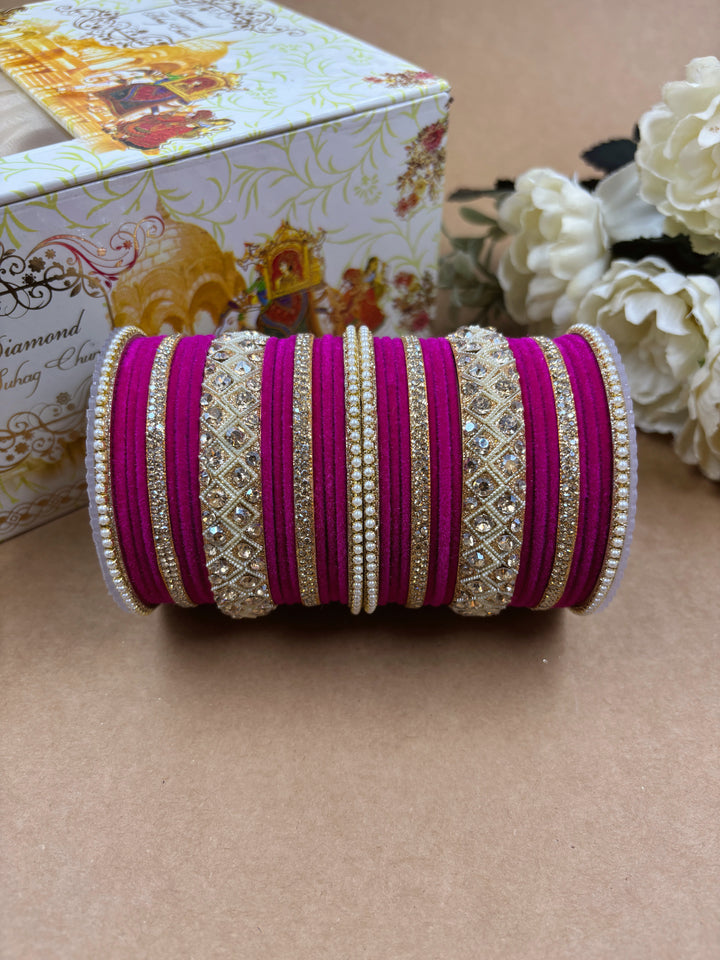 Magenta velvet bridal bangle set featuring intricate gold-toned stonework and pearl detailing, displayed beside a decorative wedding-themed box and white artificial flowers on a brown background.