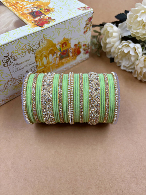 Light green velvet bridal bangle set featuring intricate gold-toned stonework and pearl detailing, displayed beside a decorative wedding-themed box and white artificial flowers on a brown background.