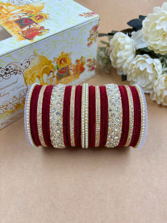 Traditional maroon velvet bridal bangle set featuring intricate gold-toned stone and pearl embellishments, displayed with a decorative wedding box and white artificial flowers on a brown background by hitesh bangles
