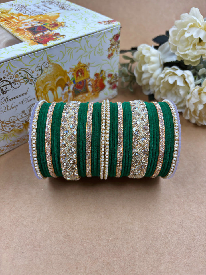 Traditional green velvet bridal bangle set featuring gold-toned bangles with intricate stone and pearl embellishments, displayed with a decorative wedding-themed box and white flowers on a brown background.