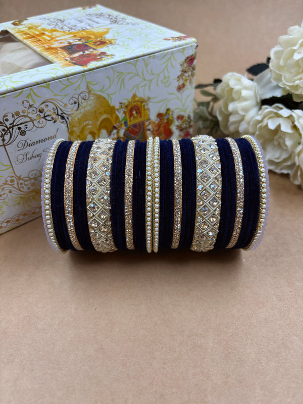 Dark blue velvet bridal bangle set adorned with gold-toned stonework and pearl accents, displayed alongside a decorative wedding-themed box and white artificial flowers on a brown background.
