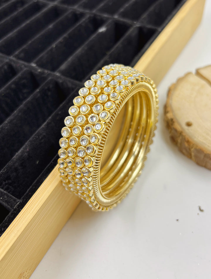 Set of gold bangles adorned with multiple rows of clear crystal stones, displayed against a black velvet tray and wooden background — ideal for weddings or festive wear.

