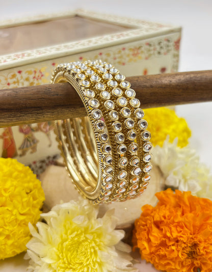 Gold bangles with white stones on a wooden stick surrounded by flowers and a decorative box by hitesh bangles