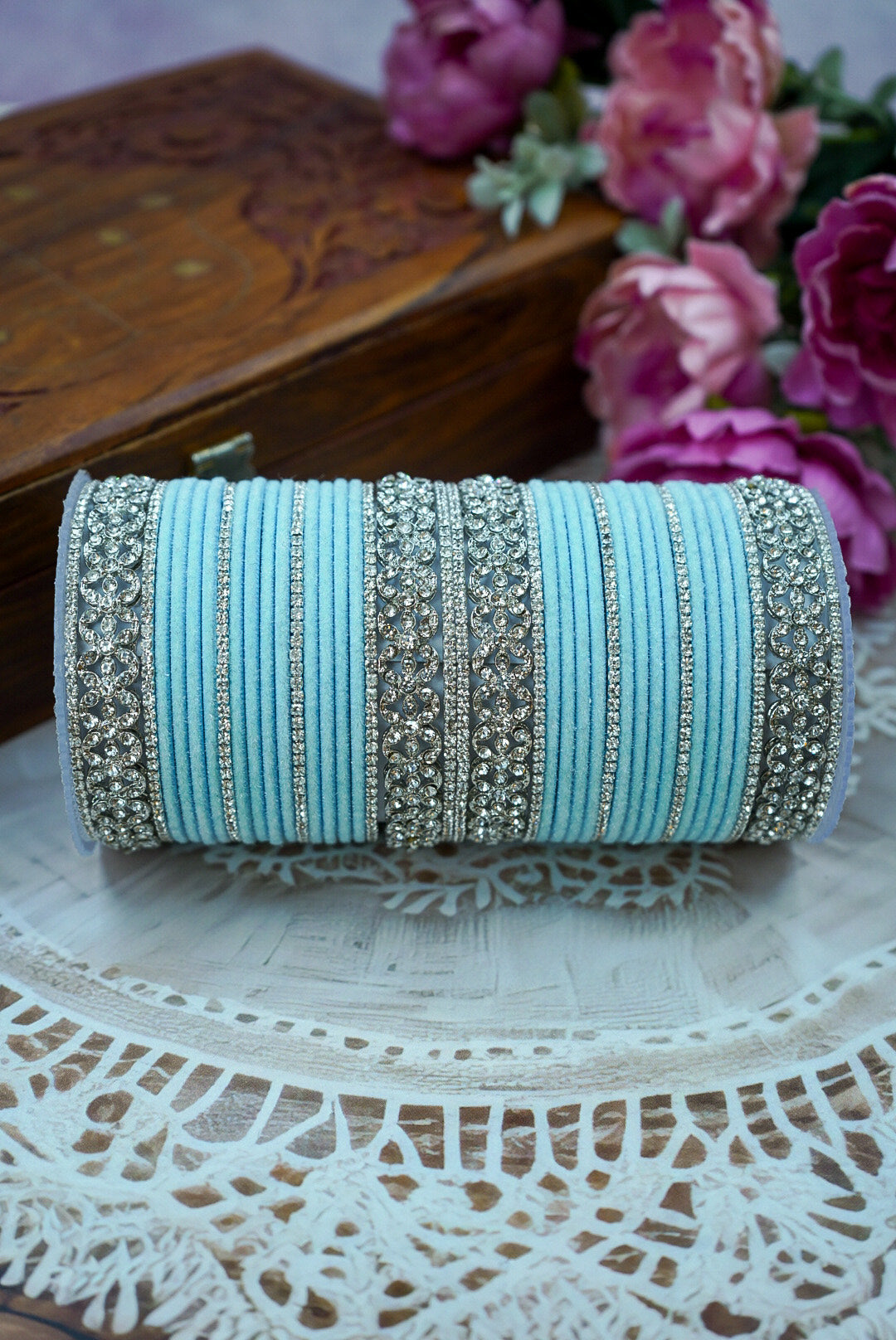 Decorative clutch with turquoise fabric and silver embellishments on a lace tablecloth with flowers in the background. by hitesh bangles