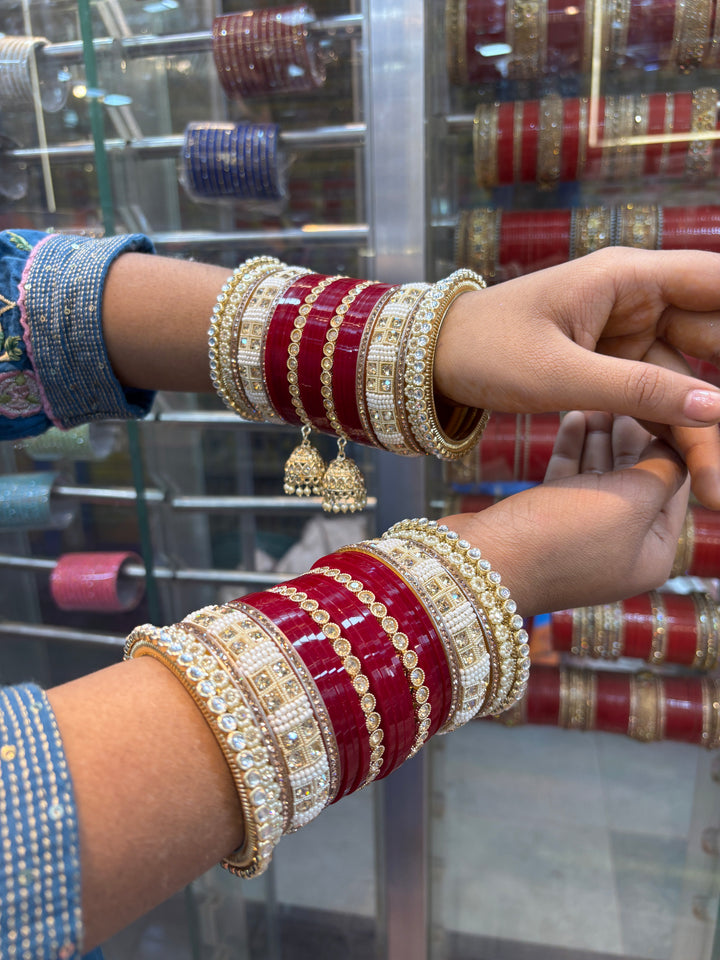 Traditional red bridal chooda set with gold and pearl detailing, featuring stone-studded kadas and jhumki hangings, worn on both arms in a jewelry store display